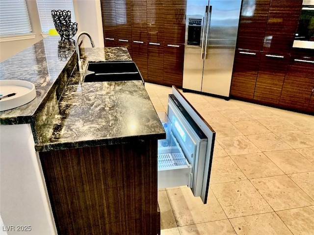 kitchen featuring a large island with sink, dark stone countertops, sink, light tile patterned flooring, and stainless steel fridge