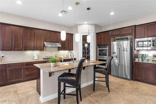 kitchen featuring a kitchen island with sink, appliances with stainless steel finishes, light stone counters, and pendant lighting