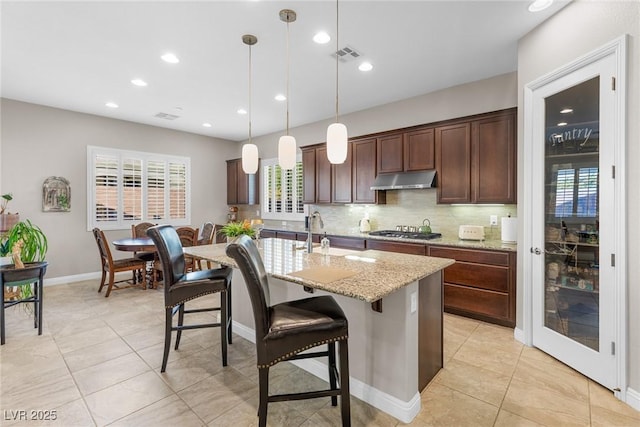 kitchen with a center island with sink, decorative light fixtures, dark brown cabinets, light stone counters, and sink