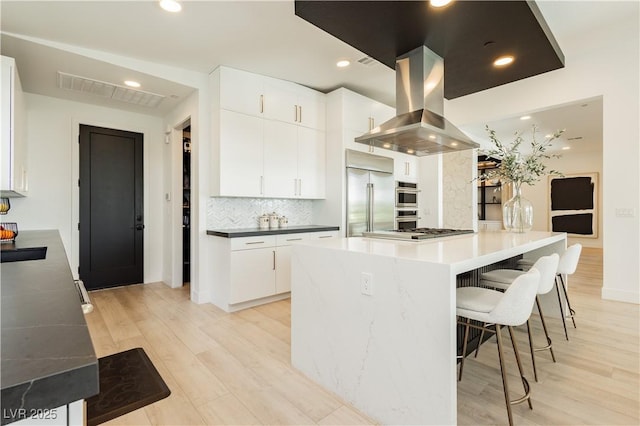 kitchen featuring white cabinets, appliances with stainless steel finishes, backsplash, island range hood, and light wood-type flooring