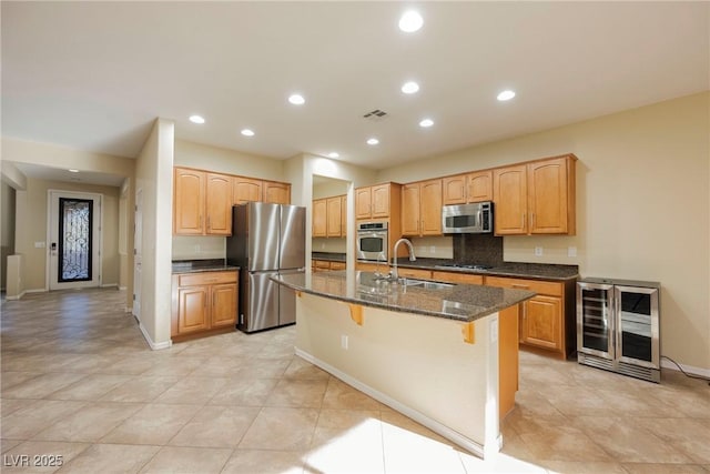 kitchen with sink, dark stone countertops, appliances with stainless steel finishes, an island with sink, and beverage cooler