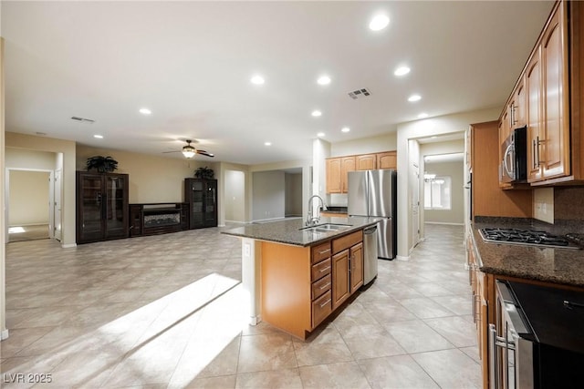 kitchen with sink, ceiling fan, appliances with stainless steel finishes, a center island with sink, and dark stone counters