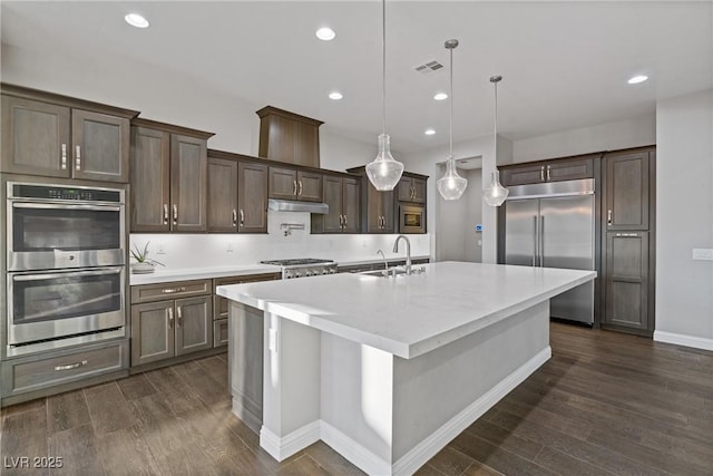 kitchen with a center island with sink, stainless steel appliances, hanging light fixtures, dark brown cabinetry, and sink