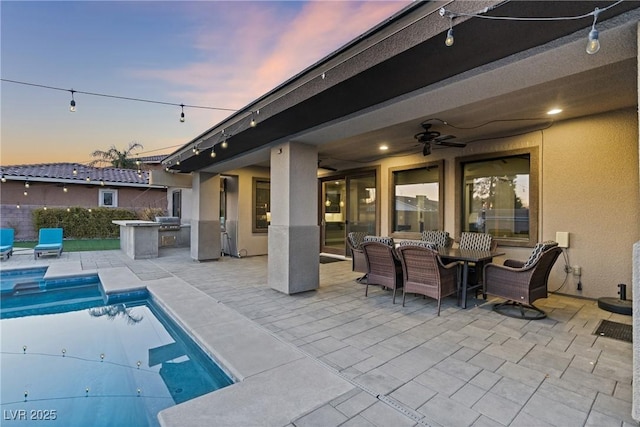 pool at dusk with ceiling fan, a grill, exterior kitchen, and a patio