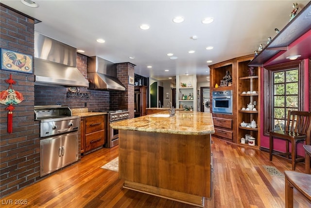 kitchen with light stone counters, appliances with stainless steel finishes, wall chimney exhaust hood, and an island with sink