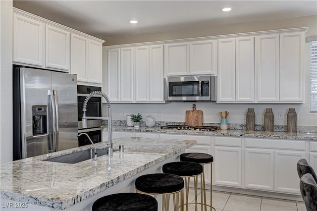 kitchen featuring white cabinetry, an island with sink, appliances with stainless steel finishes, light tile patterned flooring, and light stone counters