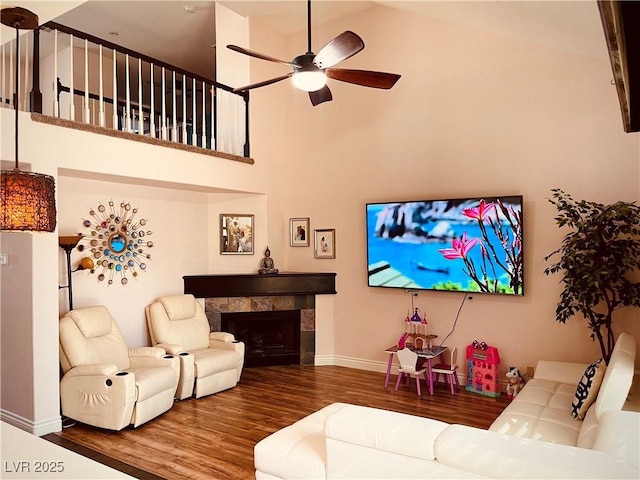 living room with ceiling fan, a tiled fireplace, a towering ceiling, and hardwood / wood-style floors