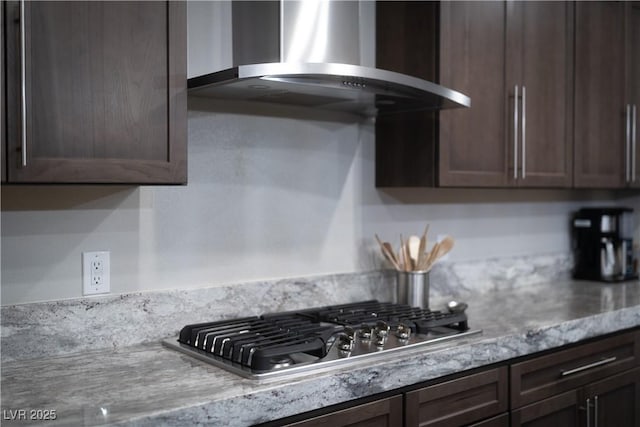 kitchen with light stone countertops, stainless steel gas stovetop, wall chimney range hood, and dark brown cabinets