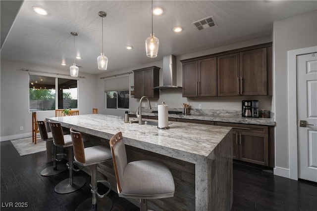 kitchen featuring pendant lighting, dark brown cabinetry, wall chimney exhaust hood, and an island with sink