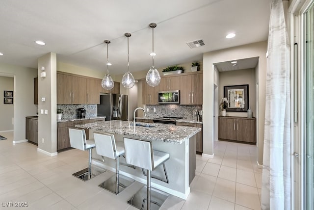 kitchen featuring decorative light fixtures, a center island with sink, sink, stainless steel appliances, and light stone counters