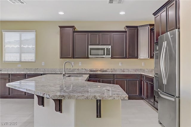 kitchen with stainless steel appliances, a kitchen breakfast bar, dark brown cabinets, light stone counters, and sink