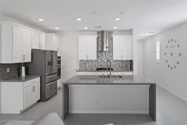 kitchen featuring wall chimney range hood, sink, a kitchen island with sink, white cabinets, and stainless steel fridge