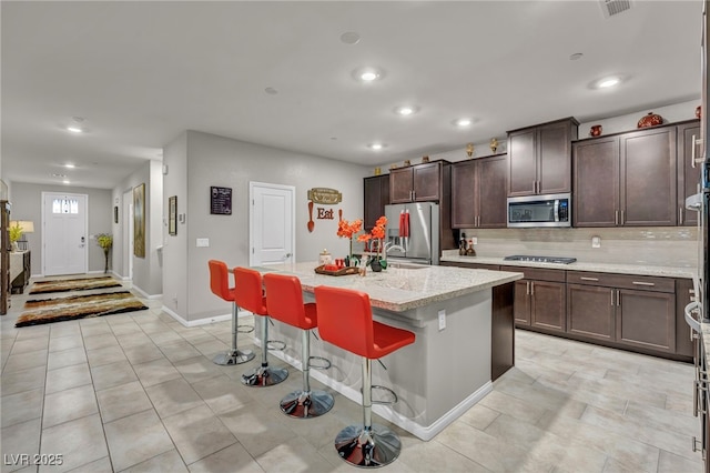 kitchen featuring a center island with sink, stainless steel appliances, a kitchen breakfast bar, dark brown cabinets, and light stone countertops