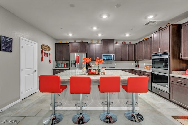 kitchen with a kitchen bar, an island with sink, stainless steel appliances, light stone counters, and dark brown cabinetry