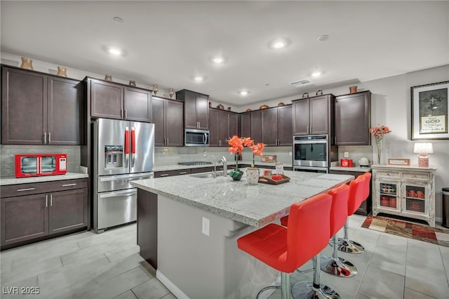 kitchen with light stone countertops, stainless steel appliances, a kitchen breakfast bar, a kitchen island with sink, and dark brown cabinets