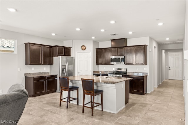 kitchen with sink, an island with sink, a breakfast bar area, stainless steel appliances, and dark brown cabinets