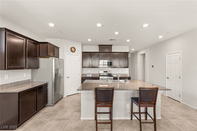 kitchen featuring an island with sink, stainless steel appliances, light stone counters, dark brown cabinetry, and sink