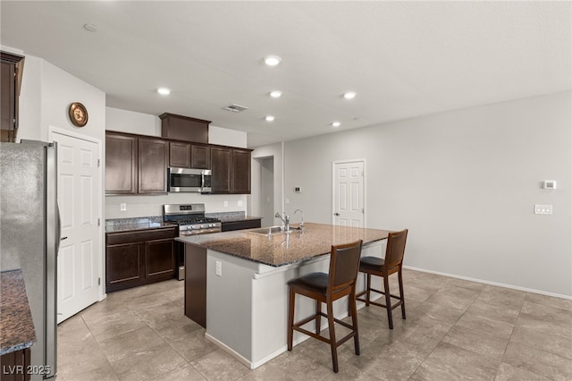 kitchen featuring a center island with sink, appliances with stainless steel finishes, dark brown cabinets, dark stone counters, and sink