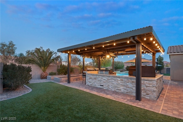 yard at dusk featuring ceiling fan, an outdoor kitchen, and a patio