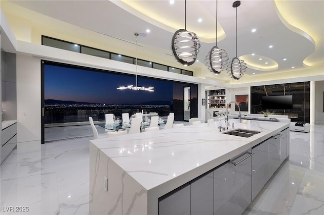 kitchen with decorative light fixtures, a large island with sink, a raised ceiling, sink, and light stone counters