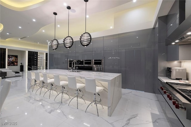 kitchen featuring light stone countertops, a large island with sink, decorative light fixtures, wall chimney range hood, and stainless steel stove