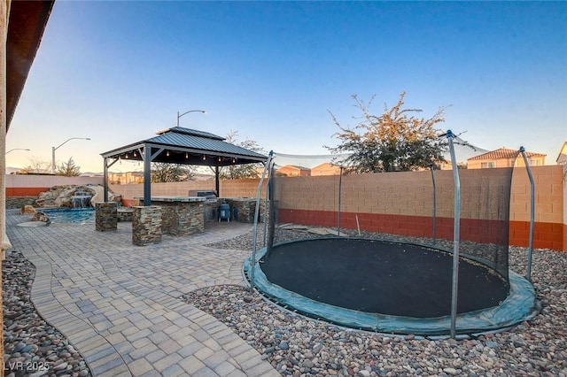 patio terrace at dusk featuring a trampoline, pool water feature, an outdoor kitchen, and a gazebo