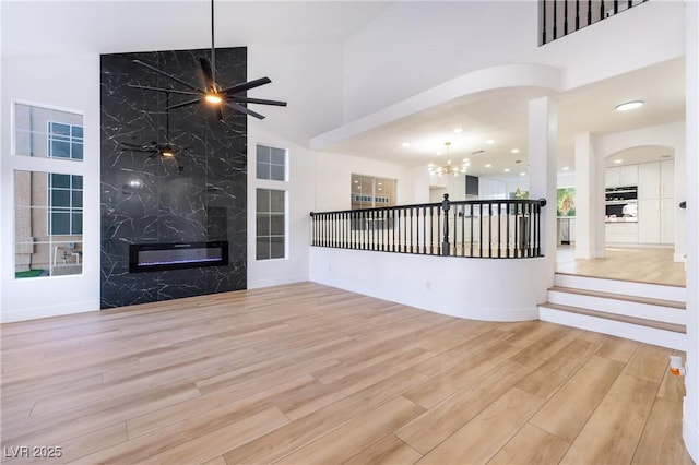 unfurnished living room with light wood-type flooring, a high end fireplace, ceiling fan with notable chandelier, and a high ceiling