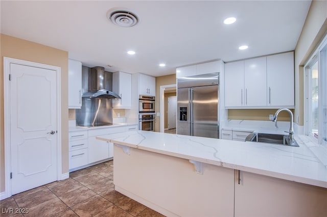 kitchen featuring wall chimney range hood, kitchen peninsula, sink, appliances with stainless steel finishes, and white cabinets