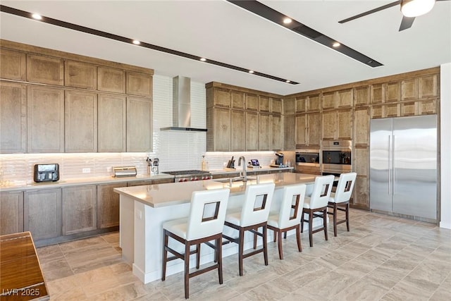 kitchen featuring stainless steel appliances, a breakfast bar, light countertops, wall chimney range hood, and a large island with sink