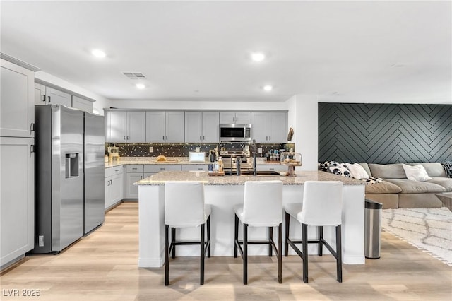 kitchen with a center island with sink, a kitchen bar, appliances with stainless steel finishes, gray cabinetry, and light stone counters