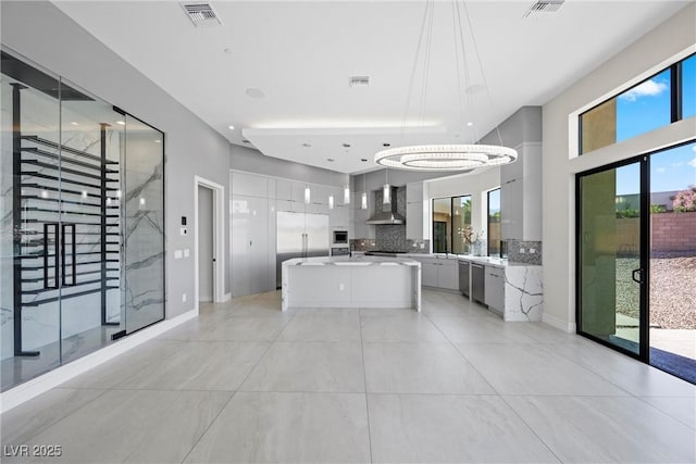 kitchen with a center island with sink, decorative backsplash, wall chimney range hood, a chandelier, and pendant lighting