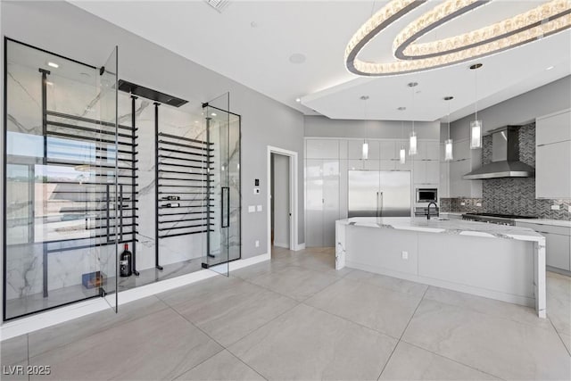 kitchen with backsplash, wall chimney range hood, a spacious island, built in appliances, and hanging light fixtures