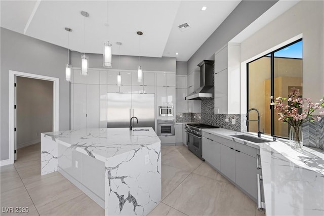 kitchen with decorative light fixtures, wall chimney range hood, built in appliances, sink, and a large island