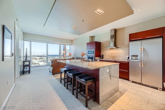 kitchen featuring a center island with sink, a breakfast bar area, stainless steel appliances, wall chimney exhaust hood, and light stone counters