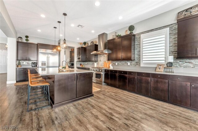 kitchen featuring high end appliances, dark brown cabinetry, hanging light fixtures, an island with sink, and wall chimney exhaust hood