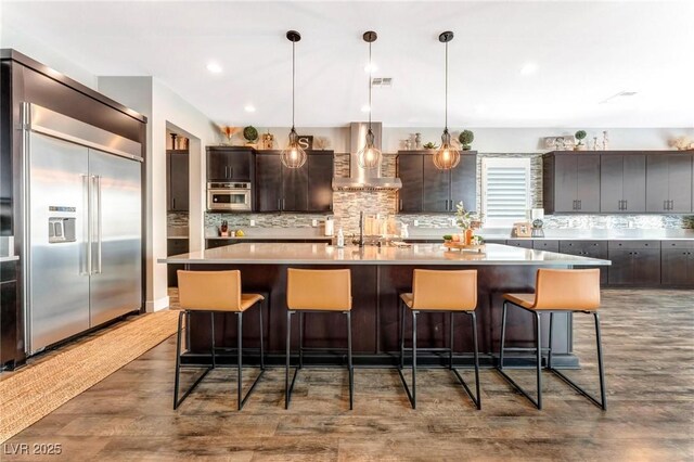 kitchen with built in refrigerator, wall chimney exhaust hood, decorative light fixtures, decorative backsplash, and a center island with sink