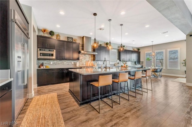 kitchen featuring an island with sink, decorative light fixtures, light wood-type flooring, dark brown cabinets, and wall chimney exhaust hood