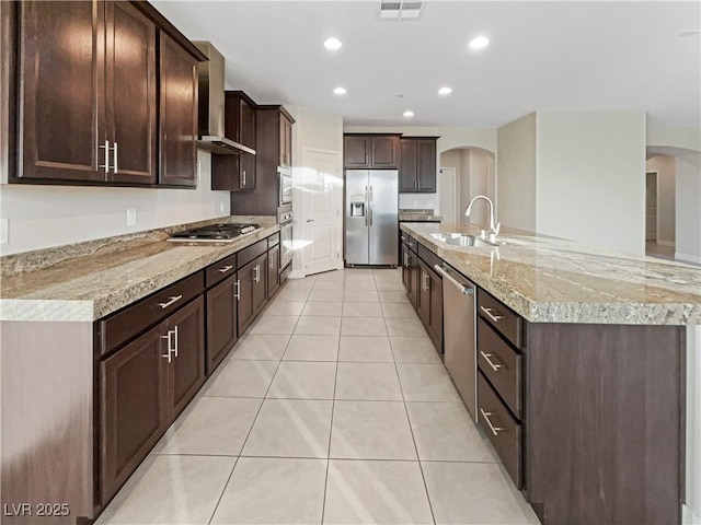 kitchen featuring light tile patterned floors, a center island with sink, stainless steel appliances, wall chimney exhaust hood, and sink