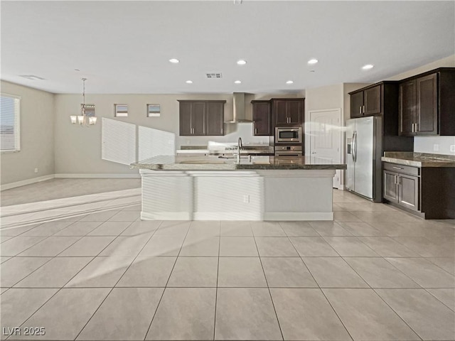 kitchen featuring light tile patterned floors, appliances with stainless steel finishes, a kitchen island with sink, dark brown cabinets, and dark stone counters