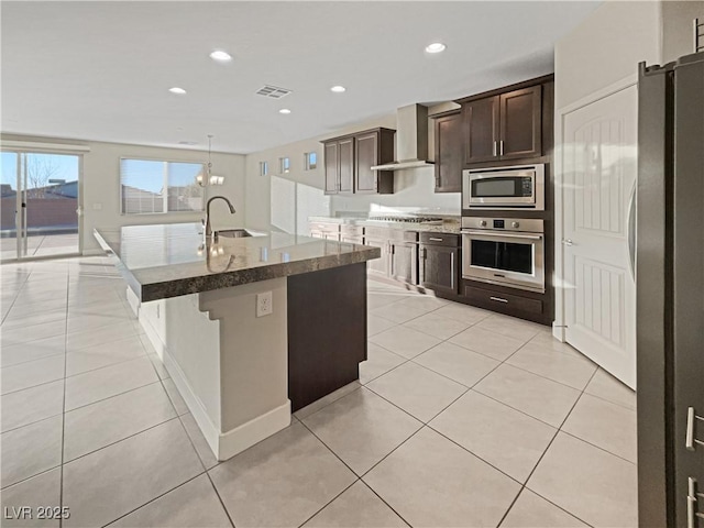 kitchen with an island with sink, stainless steel appliances, wall chimney range hood, dark brown cabinetry, and sink