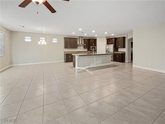 kitchen with wall chimney exhaust hood, stainless steel fridge, a kitchen island with sink, light tile patterned flooring, and ceiling fan