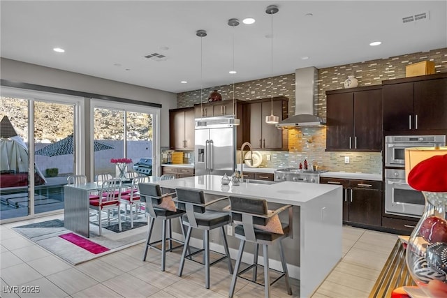 kitchen with an island with sink, pendant lighting, wall chimney exhaust hood, stainless steel built in refrigerator, and sink