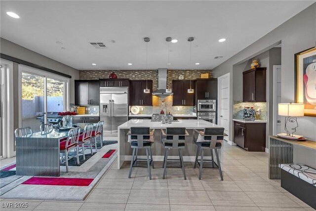 kitchen featuring a large island with sink, wall chimney range hood, dark brown cabinetry, hanging light fixtures, and appliances with stainless steel finishes