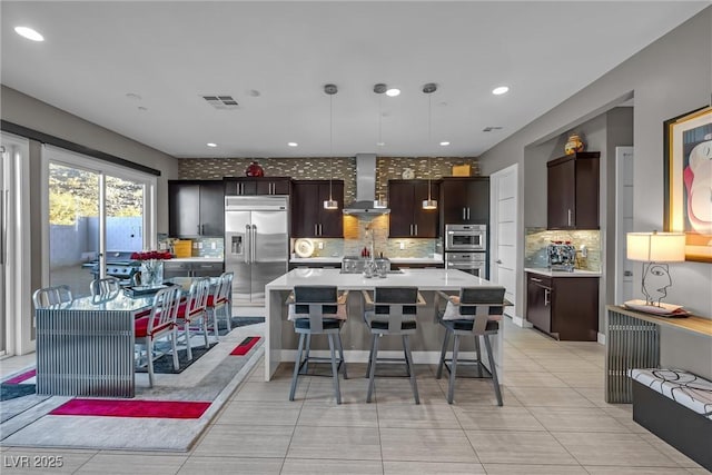 kitchen featuring appliances with stainless steel finishes, a spacious island, decorative light fixtures, wall chimney range hood, and dark brown cabinetry