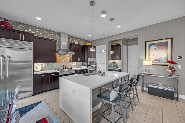 kitchen featuring appliances with stainless steel finishes, wall chimney range hood, hanging light fixtures, a kitchen island with sink, and a breakfast bar area