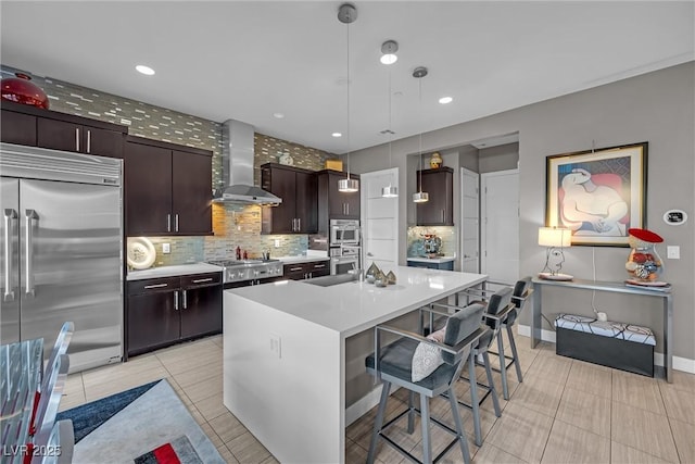 kitchen with decorative light fixtures, wall chimney range hood, a center island with sink, a kitchen breakfast bar, and stainless steel appliances