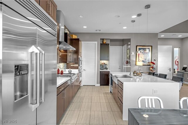 kitchen with appliances with stainless steel finishes, wall chimney exhaust hood, tasteful backsplash, sink, and a kitchen island with sink