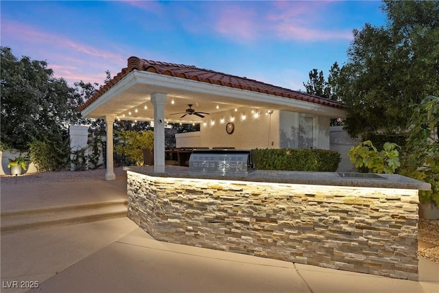 patio terrace at dusk featuring ceiling fan, an outdoor kitchen, and grilling area