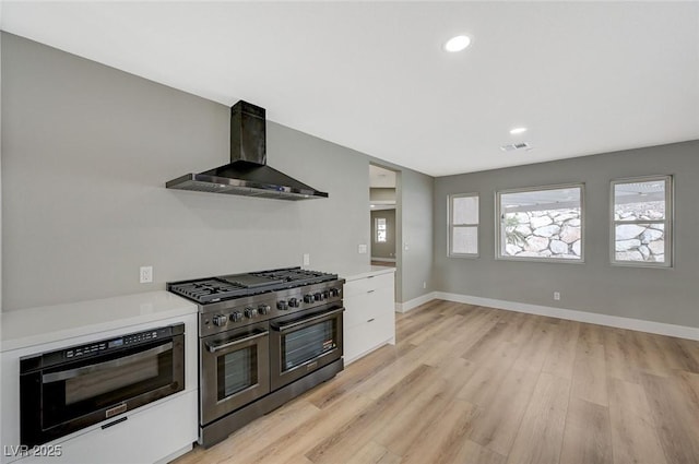 kitchen featuring white cabinetry, wall chimney range hood, range with two ovens, wall oven, and light hardwood / wood-style flooring