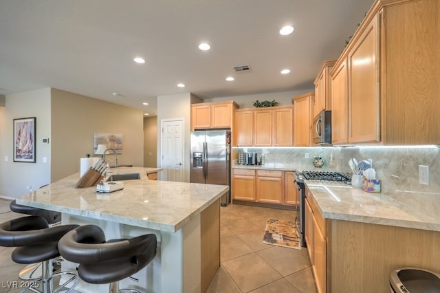 kitchen featuring decorative backsplash, an island with sink, light brown cabinets, stainless steel appliances, and light stone counters